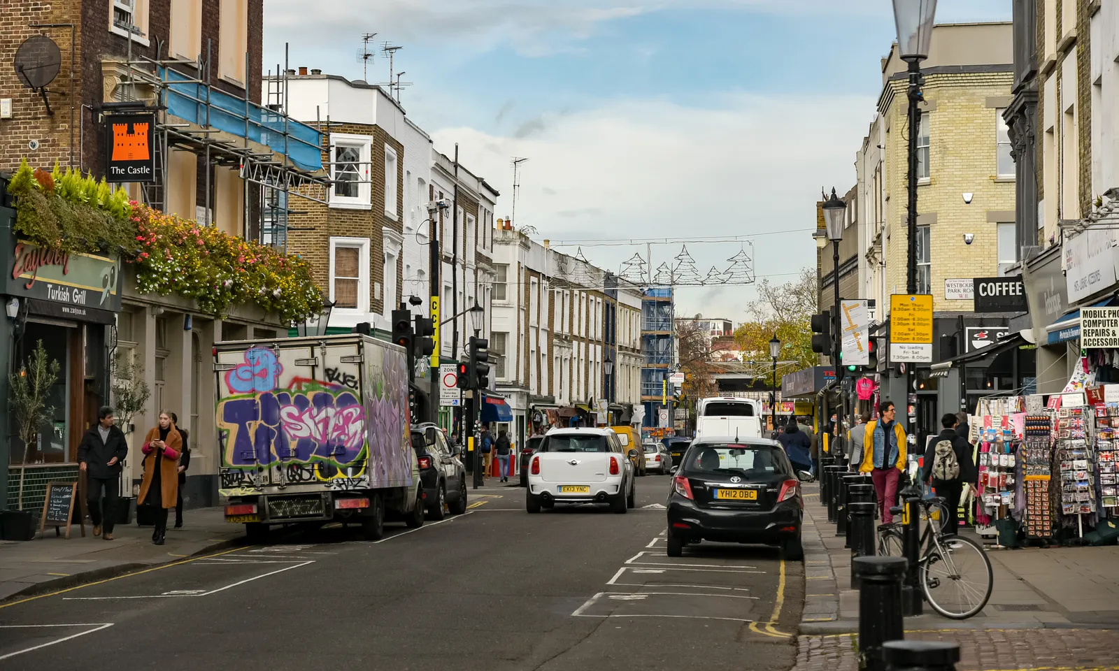A street on Portobello Road with a graffiti-covered van, parked cars and people walking past shopfronts