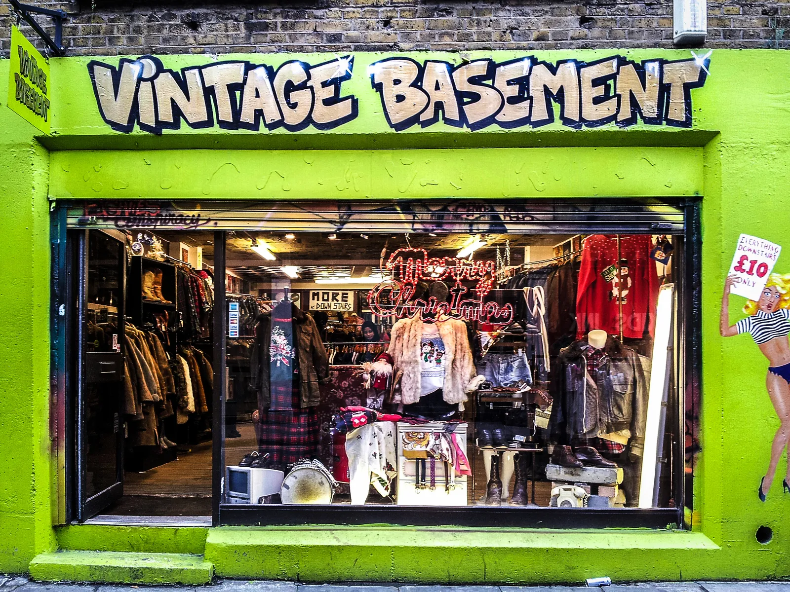 A lime-green vintage shopfront packed with rails of jackets and jumpers, with a £10 sign painted on the wall beside it
