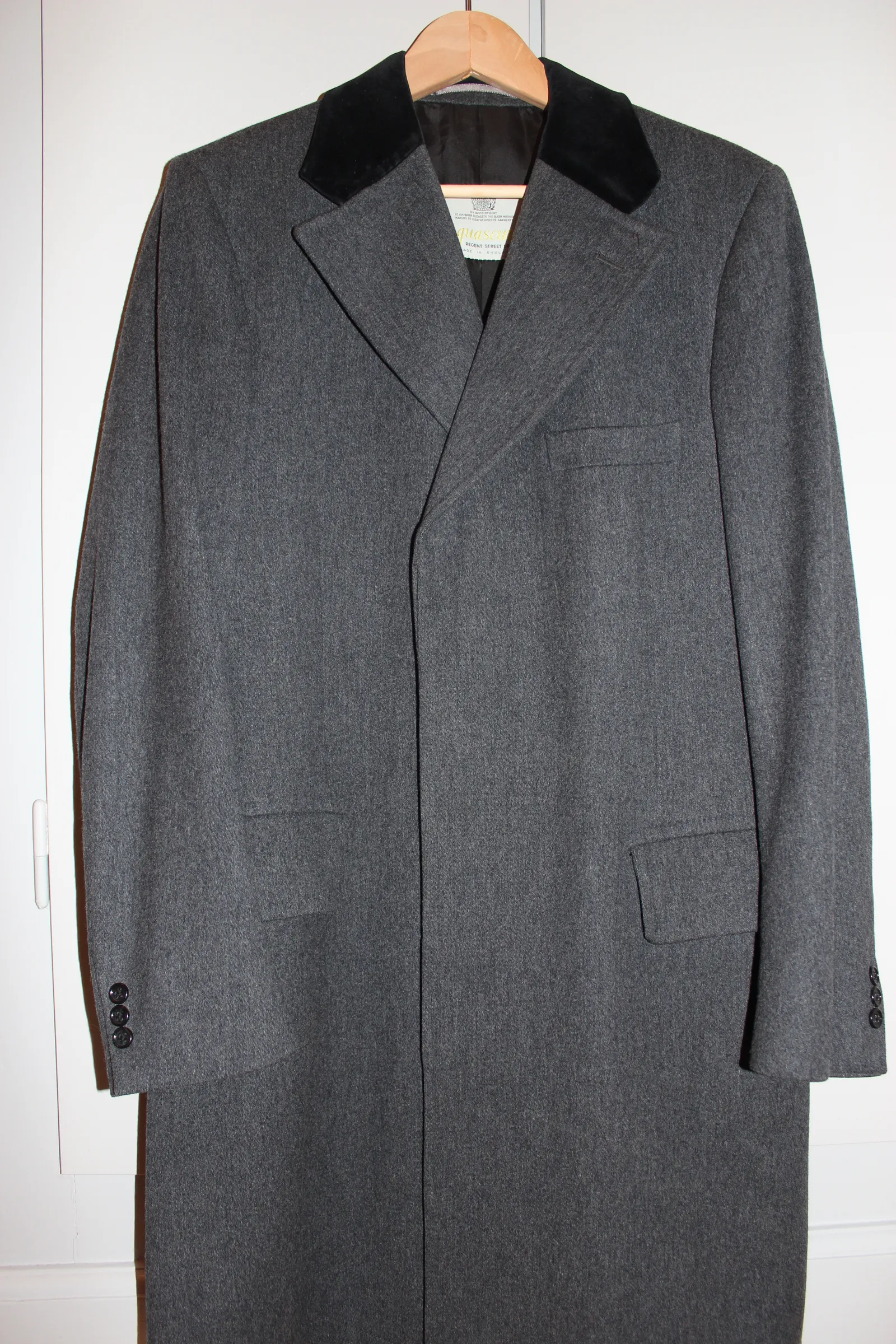 A grey wool covert coat with a dark velvet collar on a wooden hanger against a white wall