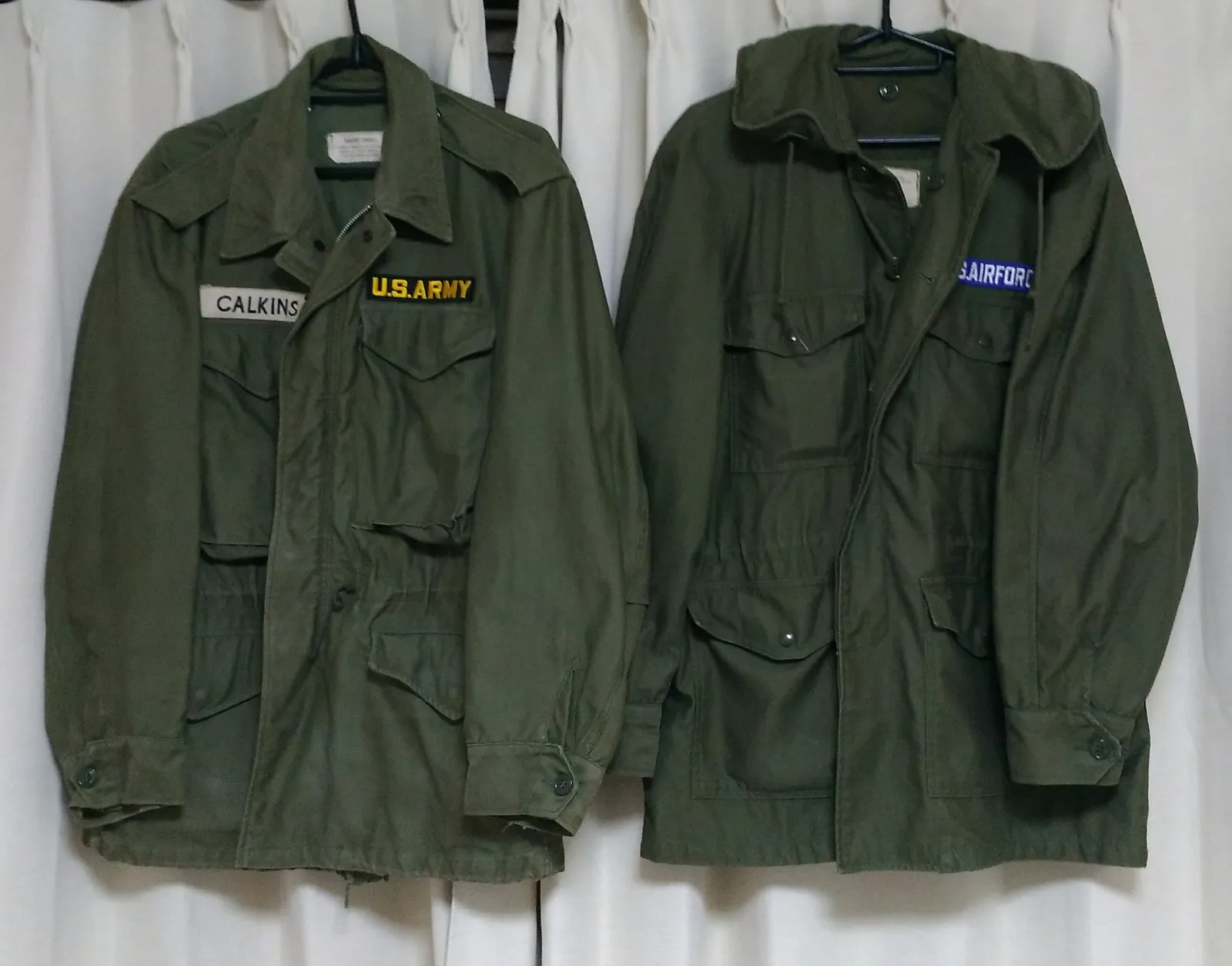 Two olive-green US military field jackets on hangers against a white curtain, one with a name tape and a U.S. Army tape, the other with a U.S. Air Force tape and its hood up