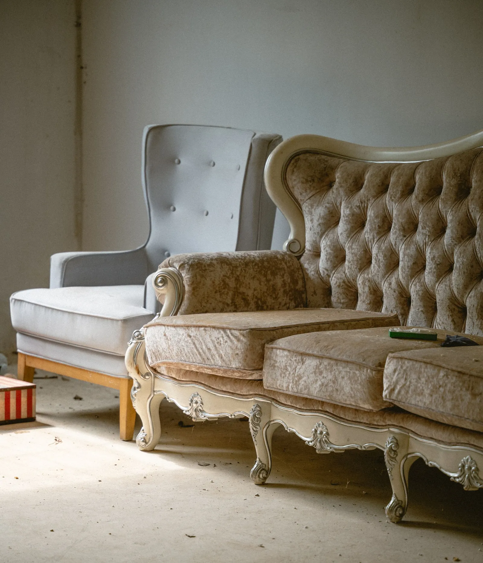 An ornate sofa in champagne crushed velvet with a carved cream frame, next to a pale grey buttoned wing chair, in a bare room with a dusty floor