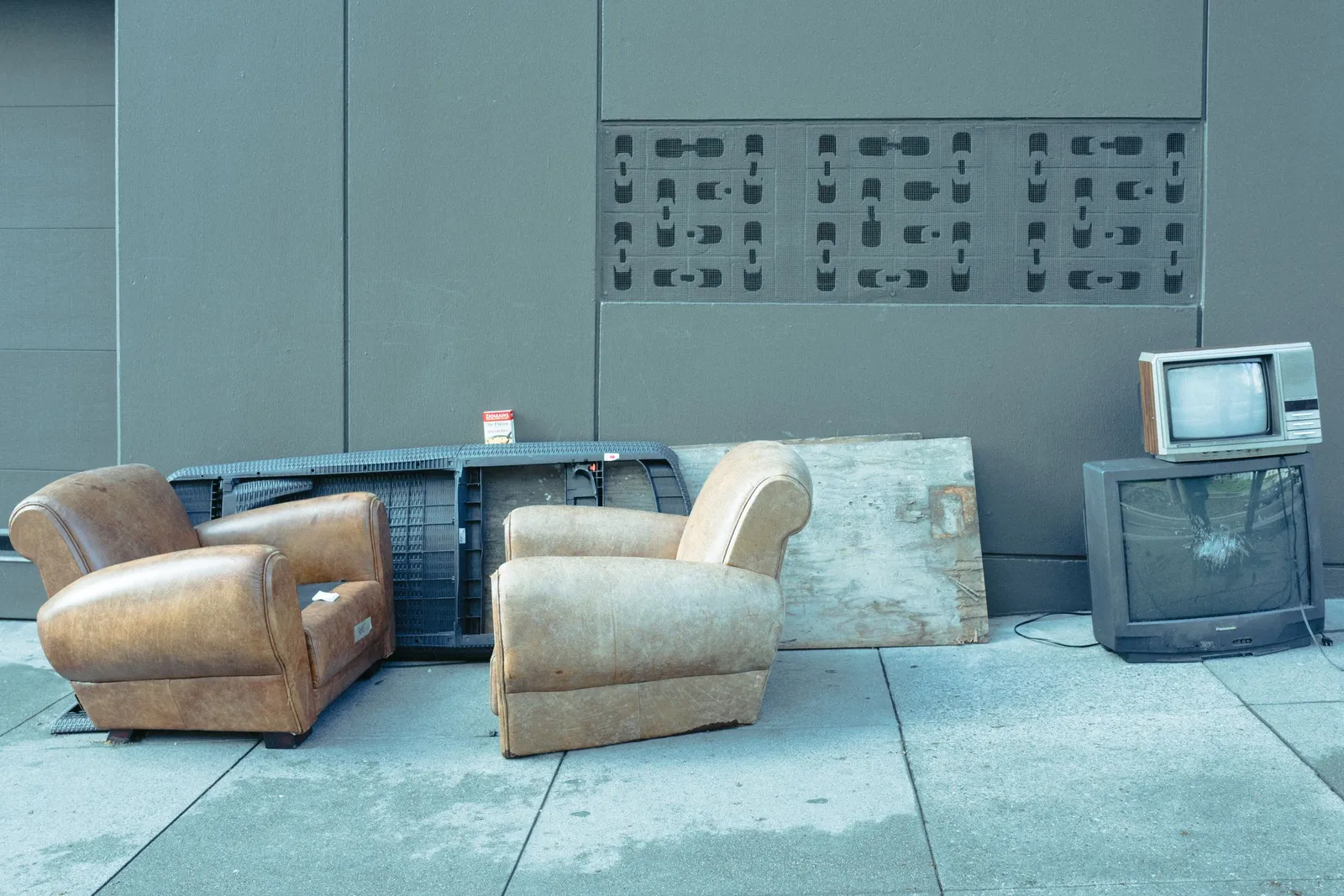 Two worn tan leather club chairs on a pavement beside a stack of old televisions, one with a cracked screen